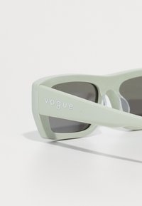 Svetlo zelene pravokotne sončne očala s temnimi lečami in vtisnjenim napisom "vogue" na roki, postavljena na belo ozadje.