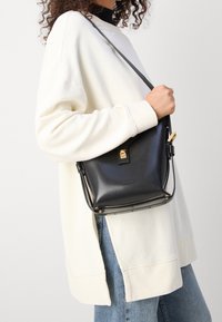 Borsa a tracolla in pelle nera con design strutturato, hardware dorato e tracolla staccabile, appoggiata contro un ampio maglione crema e un paio di jeans.
