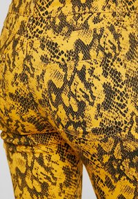 Leggings con estampado de serpiente amarillo que cuentan con un diseño texturizado con patrones negros, un ajuste ceñido y detalles de costura visibles en los bolsillos.