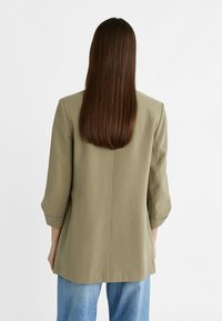 Olivgrön blazer med skräddarsydd passform, trekvartslånga ärmar och slagkrage. Buren över ljusblå denim, vilket visar en enkel och elegant design.