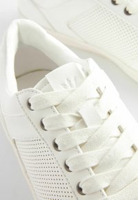 Białe skórzane sneakersy z perforowanymi bocznymi panelami, płaskimi plecionymi sznurowadłami, okrągłym noskiem oraz teksturowaną metką na języku. Widoczna gumowa podeszwa.