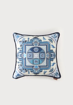 Coussin décoratif carré avec motifs géométriques et floraux bleus et blancs, bordure à pompons bleu foncé sur fond blanc.
