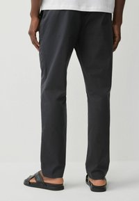 Pantalons noirs légers avec une coupe droite, présentant une texture lisse et un subtil éclat. Portés avec des sandales noires, laissant apparaître la cheville.