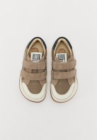 Zapatillas de ante marrón con detalles perforados, suela de goma blanca, doble correa de Velcro y forro interno beige. Presentan un acento azul marino en el talón.