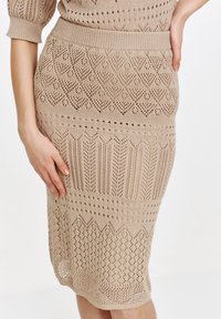 Dzianinowa spódnica ołówkowa w jasnym beżu z teksturowanym, ażurowym wzorem zawierającym geometryczne kształty oraz prążkowanym pasem w talii. Obcisły krój.