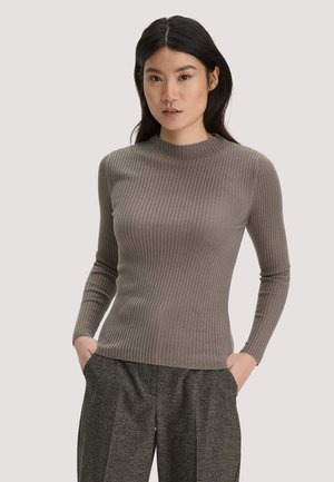 Frau mit langen schwarzen Haaren, die einen eng anliegenden braunen Rippstrickpullover und graue karierte Hose trägt, mit den Händen in den Taschen vor einem einfarbigen Hintergrund.