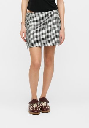 Femme portant un haut noir, une mini-jupe à carreaux noir et blanc, et des baskets marron avec des lacets crème, debout devant un fond uni.