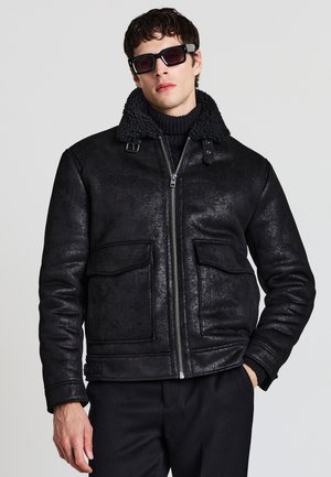 Giacca nera con colletto in shearling, chiusura con zip davanti, due tasche sul petto, texture esterna liscia, con una vestibilità aderente e un design moderno.