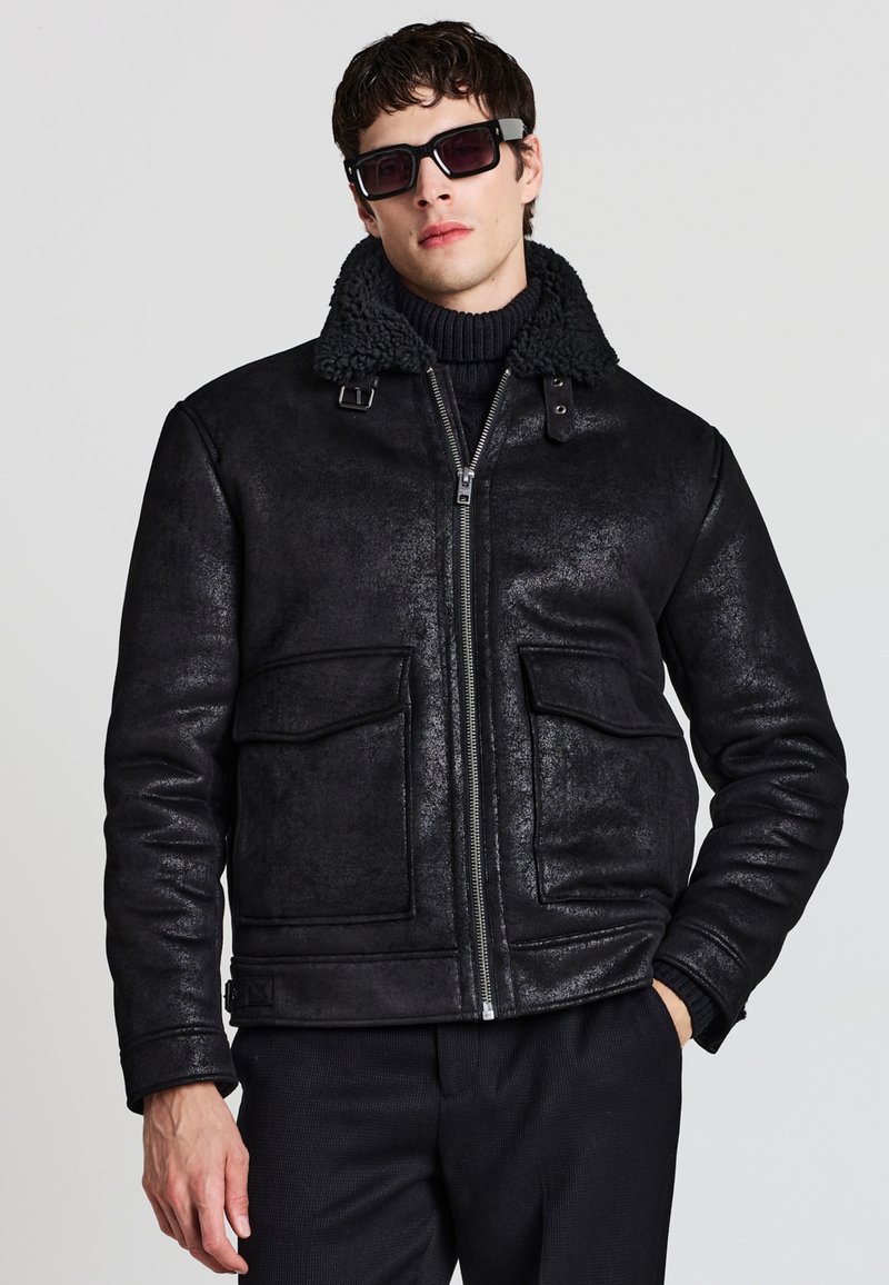 Giacca nera con colletto in shearling, chiusura con zip davanti, due tasche sul petto, texture esterna liscia, con una vestibilità aderente e un design moderno.