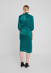 Teal satinklänning i midi-längd med hög krage, puffärmar och en dold dragkedja i ryggen, med figursydd midja och något utsvängd nederdel. Svarta ankelskängor.