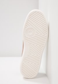 Semelle de chaussure en caoutchouc blanche avec motif géométrique de la bande de roulement appuyée contre un mur blanc sur une surface blanche.