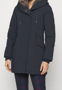 Marinblå parka med foder av konstpäls i huva, dragkedja och tryckknappar, ribbade ärmslut och två framfickor. Har en dekorativ logotypdetalj.