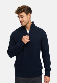 Marineblå ribstrikket sweater med høj krave og halvlynlås; fremstillet af blødt materiale med lange ærmer og en afslappet pasform.