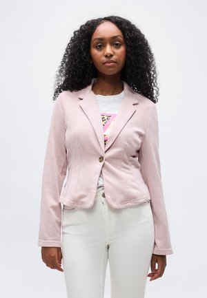 Ung kvinde med krøllet hår iført en lyserød knapblazer over en hvid t-shirt med grafik og hvide bukser, stående mod en ensfarvet baggrund.