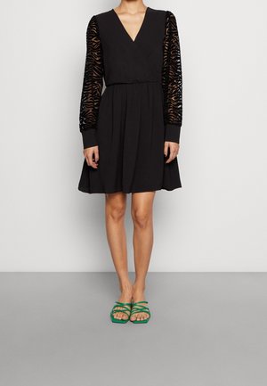 Black knee-length dress with V-neck and sheer patterned long sleeves, paired with green strappy open-toe sandals on gray floor.