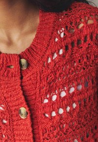 Close-up van een persoon in een rode grofgebreide cardigan met grote knopen over een witte ribbeltop.