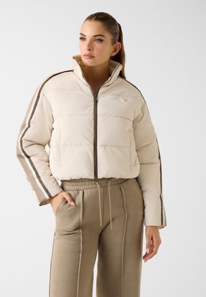 Femme aux longs cheveux attachés en queue de cheval, portant une doudoune crème avec des manches rayées et un pantalon beige à cordon, debout avec une main dans la poche.