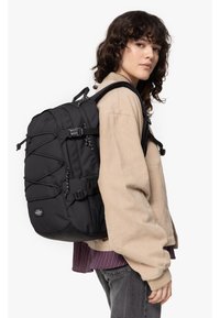 Mochila negra con acabado texturizado, correas ajustables, cuerdas elásticas externas, múltiples bolsillos y un logo redondo en la parte frontal.