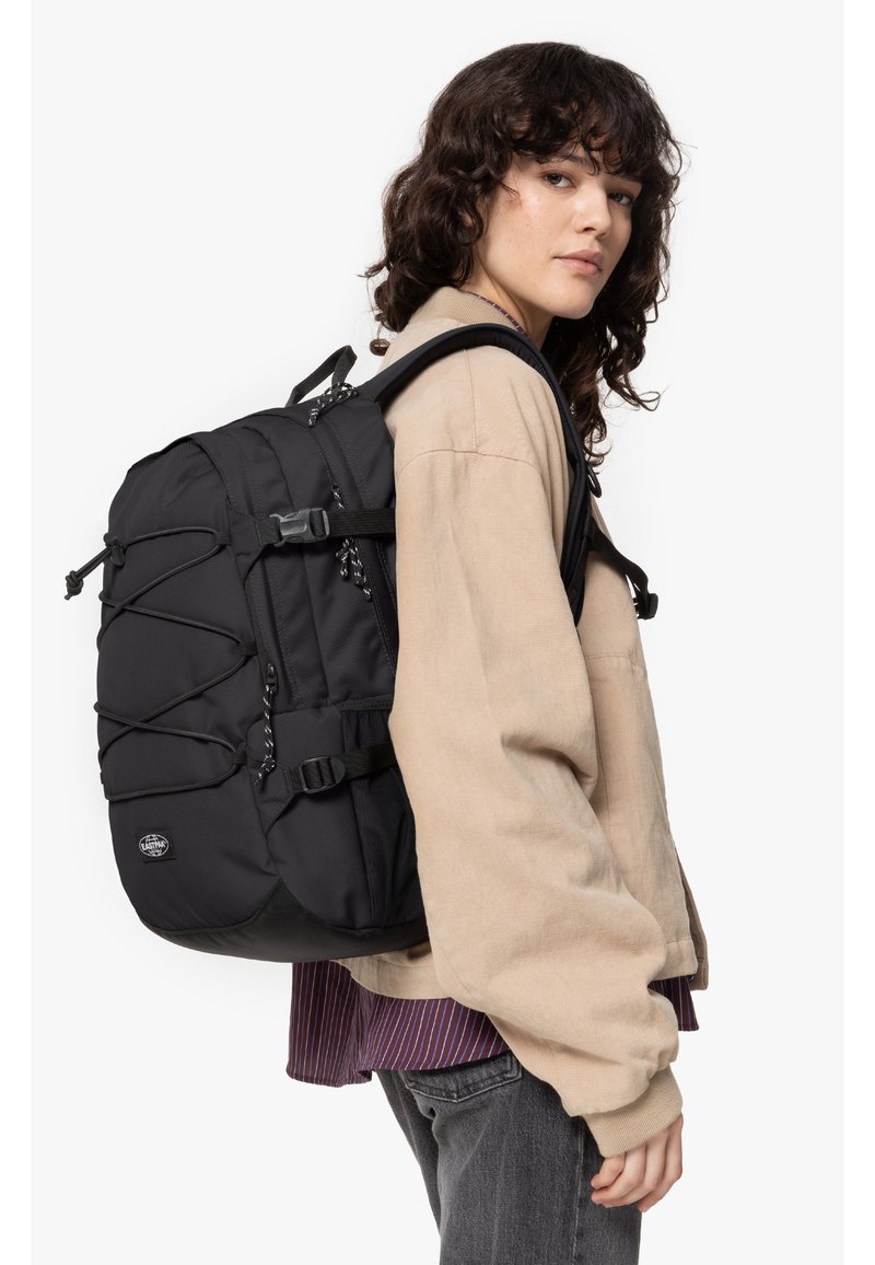 Mochila negra con acabado texturizado, correas ajustables, cuerdas elásticas externas, múltiples bolsillos y un logo redondo en la parte frontal.