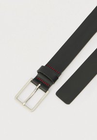 Juodas odinis diržas su sidabrine stačiakampio formos sagtimi ir raudonos siūlės detalė netoli sagties ant šviesaus fono.