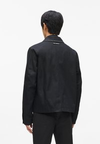 Melnā jakā ar asu apkakli, rotāta ar sudraba kniedēm ap apkakli, izgatavota no gludas auduma, ar taisnu piegriezumu un garām piedurknēm.