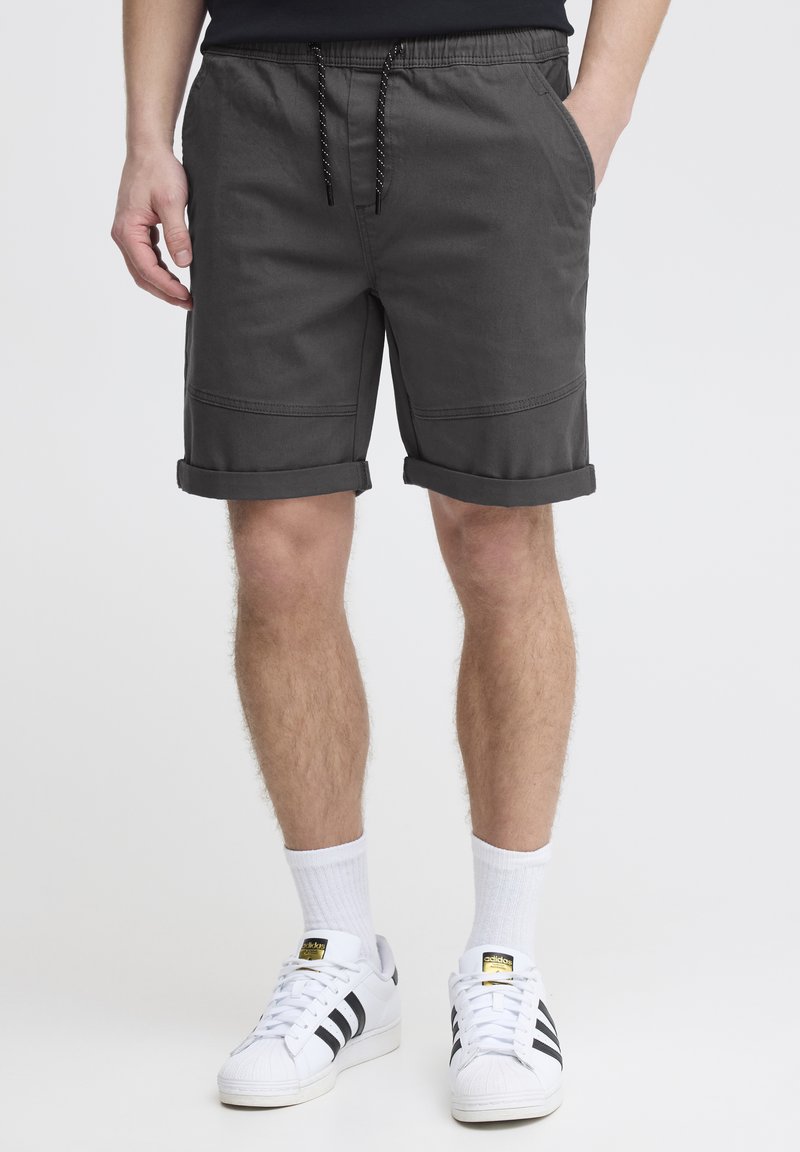 Graue Baumwollshorts mit elastischem Bund, Kordelzug, umgeschlagenen Säumen und Seitentaschen. Getragen mit weißen Turnschuhen und Socken.