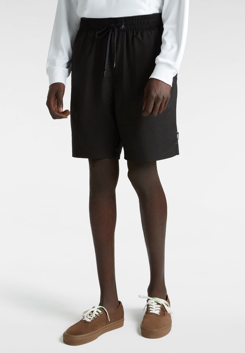 Zwarte zwemshorts met een elastische tailleband en trekkoord, voorzien van een zijlogo patch. Gepaard met bruine suède sneakers en witte veters.