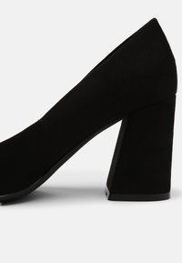 Chaussure à talon haut en suede noire avec une forme géométrique, un talon épais, une texture lisse et des caractéristiques de design minimalistes.