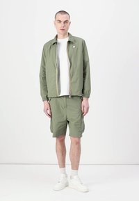 Jachetă ușoară de culoare verde oliv, cu fermoar, asortată cu pantaloni scurți în aceeași nuanță. Cămașă albă pe dedesubt și pantofi sport albi. Design minimalist.