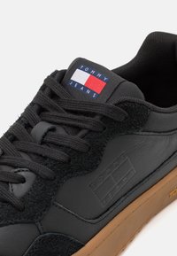 Baskets noires avec lacets noirs, semelle marron et patch logo Tommy Jeans bleu, blanc et rouge sur la languette.