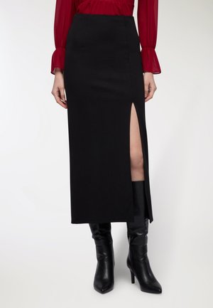 Donna che indossa una gonna midi nera con uno spacco frontale, abbinata a stivali neri con tacco e a un top rosso a maniche lunghe con polsini a campana.