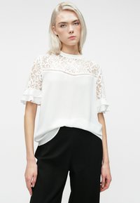 Νεαρή γυναίκα με ανοιχτόξανθα μαλλιά, φορώντας λευκή δαντελένια μπλούζα με κοντά μανίκια και μαύρο παντελόνι, που στέκεται μπροστά σε ανοιχτό φόντο.