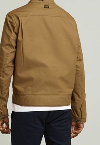 Karamellfärgad bomullsjacka med en enkel design, som har krage, knäppta manschetter och en etikett på baksidan. Bärs över en vit skjorta.