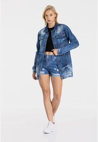 Denim jack en shorts in blauwe tinten, met versleten details en knoopaccenten. Model draagt een zwarte top en witte sneakers.