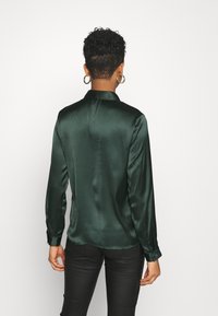 Blusa in raso verde scuro con maniche lunghe, colletto e polsini con bottoni. Tessuto liscio e design leggermente aderente.