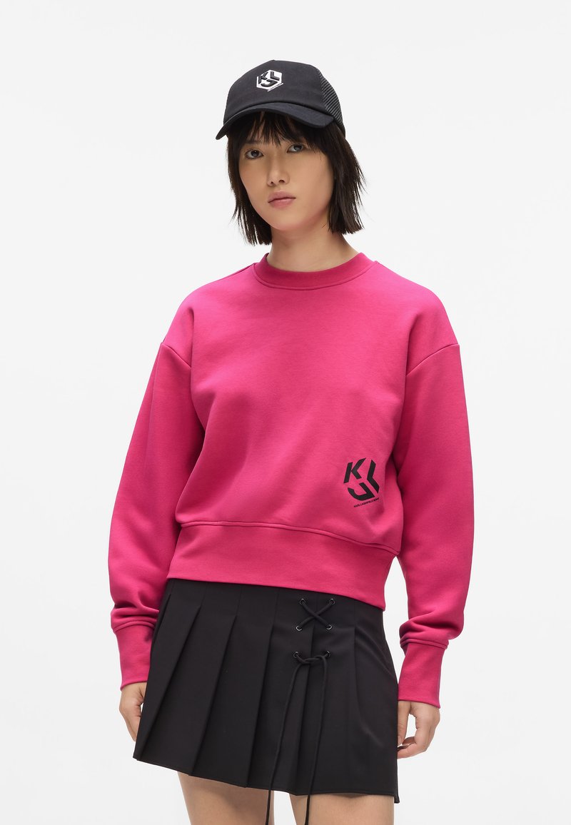 Felpa fucsia con colletto e polsini a coste, con un logo nero sul davanti. Abbinata a una gonna nera plissettata con dettagli in pizzo.