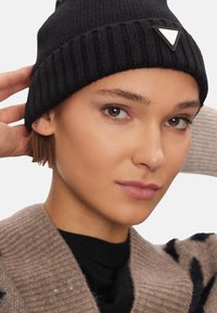 Berretta nera in maglia con texture a coste e una toppa con logo triangolare. Modello che indossa un cardigan beige con un motivo a fantasia.