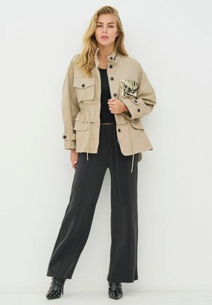 Beež jakk, millel on mitu taskut ja metallilise mustriga plaastriga, kombineeritud mustade laia säärega pükste ja naelutatud mustade pahkluu saabastega.