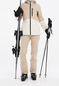Personne en tenue de ski beige tenant des skis et des bâtons, portant des gants noirs et des bottes de ski sur un fond blanc.