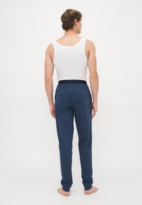Man staat blootvoets met zijn rug naar de camera, gekleed in een witte mouwloze top en marineblauwe sweatpants, tegen een effen witte achtergrond.
