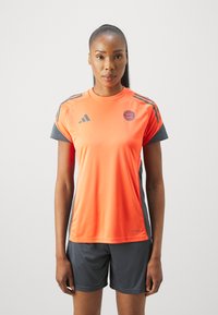 Πορτοκαλί αθλητικό μπλουζάκι με γκρι λεπτομέρειες, με κοντά μανίκια και λογότυπο Adidas. Συνδυασμένο με γκρι σορτσάκι. Λείος, αναπνεύσιμος ύφασμα.