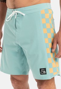 Svietlo modré plavky s bočným panelom, ktorý má striedajúce sa žlté a svetlo modré štvorce. Nastaviteľný pás so šnúrkou a logo nášivka.