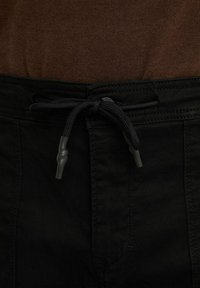 Pantalon noir avec un cordon noir noué et une braguette zippée noire, porté avec un haut en maille marron rentré à l'intérieur.