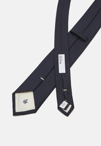 Marineblå slips laget av teksturert stoff, med en hvit underside med brodert logo, og et vedlagt omsorgsetikett i den ene enden.
