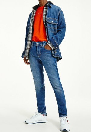 Man draagt blauwe spijkerbroek, witte sneakers, feloranje shirt, gelaagd met een geruit overhemd en een blauwe spijkerjas, staat met één hand in de zak.