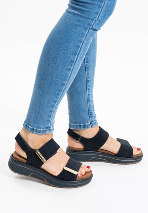 Sandali in suedè blu navy con suola piatta, dotati di due cinturini larghi, dettagli in metallo dorato e design con schiena aperta.
