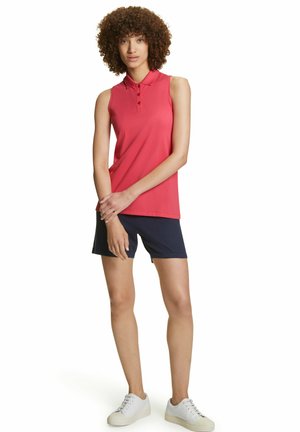 Rotes ärmelloses Poloshirt mit Kragen und geknöpftem Schlitz, kombiniert mit marineblauen Shorts. Trägt weiße Sneakers. Glatte Stofftextur.
