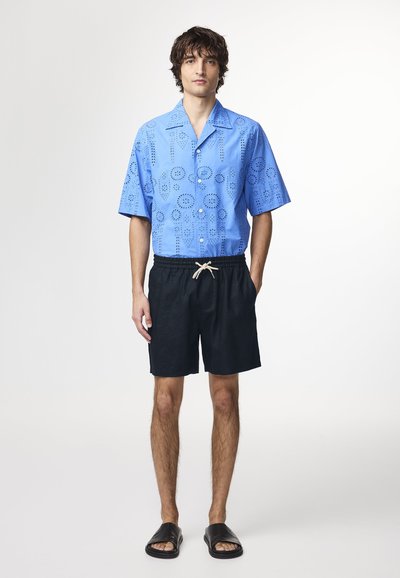 Camicia azzurra a maniche corte con motivi ad occhiello, abbinata a pantaloni corti neri con coulisse e sandali neri scivolabili su uno sfondo neutro.