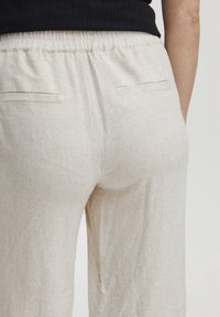 Detailní záběr na zadní část světle béžových lněných kalhot s elastickým pasem a dvěma horizontálními klínovými kapsami, které jsou nošeny s černým topem.