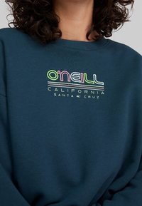 Felpa blu scuro con scollatura a girocollo, caratterizzata da una grafica in stile retrò dai colori neon che recita "O'Neill California Santa Cruz". Tessuto morbido.